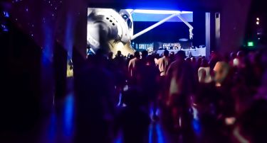 Exiting the vestibule, after the curtain they had been projecting onto has been raised, entering the main exhibit hall containing the Space Shuttle Atlantis.